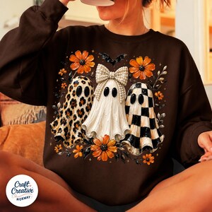 May include: A dark brown long-sleeve sweatshirt featuring a graphic print of three ghosts. The left ghost has a leopard print pattern, the center ghost wears a large pearl-embellished bow, and the right ghost has a black and white checkerboard pattern.