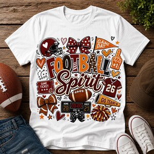 Op de afbeelding: Een wit t-shirt met een kleurrijke afbeelding met de tekst "FOOTBALL SPIRIT" omringd door illustraties van een footballhelm, stippen strikken, een wimpel, een schuimvinger, een wedstrijdkaartje, een football, een scorebord en een megafoon.