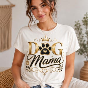 May include: Cream-colored t-shirt with the text "DOG Mama" in gold and black. The design includes a paw print, crown, and decorative flourishes. The shirt is knotted at the waist and worn with blue jeans.