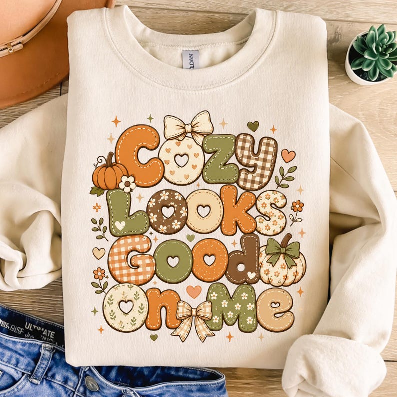 May include: A cream-colored crewneck sweatshirt featuring a multi-colored graphic print with the text "Cozy Looks Good On Me." The design includes various autumn-themed patterns, pumpkins, bows, hearts, and small flowers in shades of orange, green, and brown.