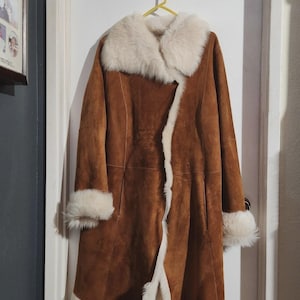 May include: A brown suede coat with a cream-colored fur collar, cuffs, and trim. The coat is hanging on a yellow hanger against a white door. The coat is a long, knee-length style.