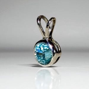 May include: A silver pendant with a round, light blue gemstone. The gemstone is set in a bezel setting and has a sparkling, textured appearance. The pendant has a loop at the top for a chain.