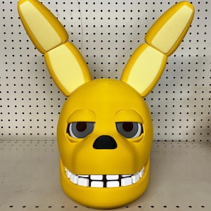 May include: A yellow rabbit head mask with large, upright ears. The mask has a wide, toothy grin with white teeth, a black nose, and dark eyes. The ears are segmented with a lighter yellow interior.