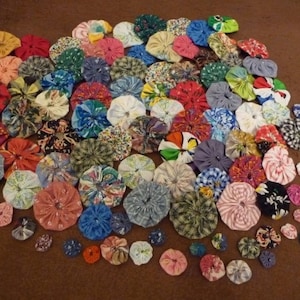 May include: A collection of colorful, handmade fabric yo-yos in various sizes. The yo-yos are made from a variety of patterned fabrics, including floral, plaid, and solid colors. They are arranged on a brown surface.