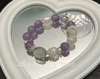 May include: A bracelet featuring alternating purple and clear beads, accented with silver-toned metal. The bracelet is displayed on a heart-shaped mirror within a white frame. The text "Aura Blossom Crystals" is visible on the mirror.