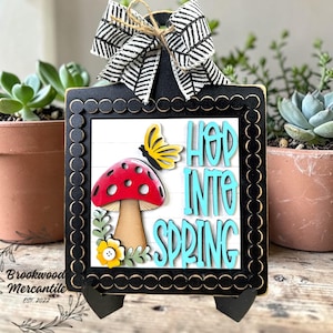 May include: A black framed sign with the words "HOP INTO SPRING" in turquoise letters. The sign features a red mushroom, yellow butterfly, and a black and white patterned bow. The sign is supported by a black stand.