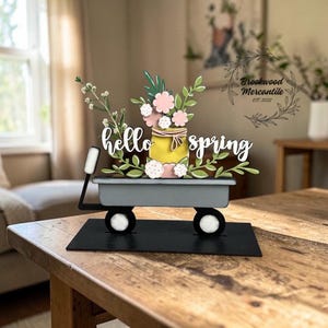 May include: A decorative spring-themed wagon with the words "hello spring". The wagon bed is gray, and it holds a yellow jar with pink and white flowers and green leaves. The wagon sits on a black base with white wheels.