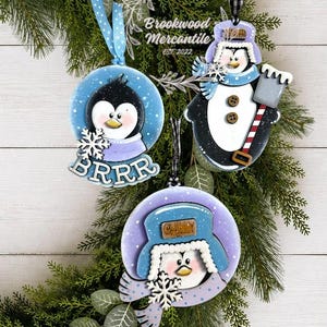May include: Three hanging ornaments featuring penguin designs. One is a round blue ornament with a penguin and the text BRRR. Another is a penguin wearing a blue hat and scarf with a shovel. The third is a penguin in a blue trapper hat.