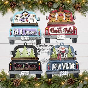 May include: Four wooden truck ornaments with holiday themes. Top left: light blue truck with snowmen and "let it snow 2026". Top right: red truck with reindeer and "North Pole 2026". Bottom left: black truck with trees and "Tree Farm Christmas". Bottom right: blue truck with snowman and "HELLO WINTER BRRR".