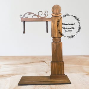 May include: A wooden sign holder with a dark brown finish. The sign holder has a base, a vertical post with decorative elements, and an arm with a scrolled design. The text "Brookwood Mercantile Est.2022" is in a circular wreath.