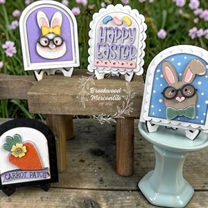 Pode incluir: Uma cole&ccedil;&atilde;o de placas de madeira com tema de P&aacute;scoa. As placas apresentam desenhos de coelhos e cenouras, com as palavras "Happy Easter" e "Carrot Patch". As placas s&atilde;o em v&aacute;rias cores, incluindo branco, roxo, azul e laranja. As placas s&atilde;o exibidas em pequenos cavaletes.