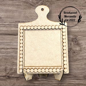 May include: A light-colored wooden cutting board with a scalloped edge and a handle. The board has a square center and is supported by two angled feet. The text "Brookwood Mercantile Est.2022" is in a circle on the right side.