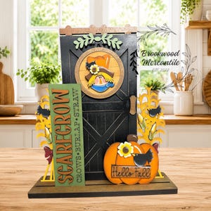 May include: A decorative wooden tabletop display featuring a black barn door with a scarecrow face in a circular frame, a green sign reading SCARECROW CROWS BURLAP STRAW, and an orange pumpkin with the text Hello Fall and a black bird.