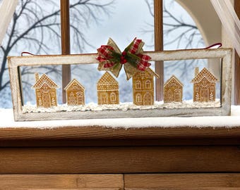 May include: A decorative window frame featuring miniature gingerbread houses with white icing details. A red and green plaid bow adorns the top, adding a festive touch. The houses sit on a bed of faux snow, creating a winter scene.