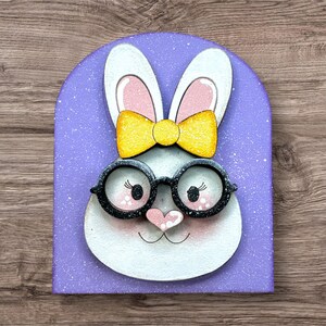 Pode incluir: Uma decora&ccedil;&atilde;o de coelho da P&aacute;scoa caprichosa. O coelho tem pelo branco, orelhas forradas de rosa e um la&ccedil;o amarelo. Ele usa &oacute;culos com arma&ccedil;&atilde;o preta e tem um nariz em forma de cora&ccedil;&atilde;o rosa. O fundo &eacute; um arco de lavanda com manchas brancas.