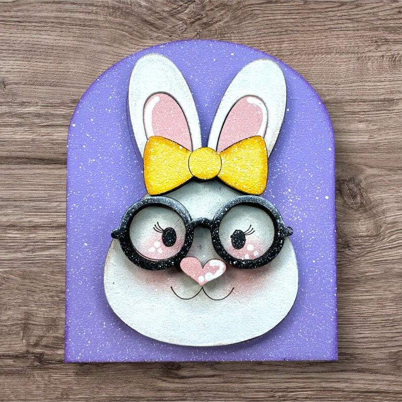 Pode incluir: Uma decora&ccedil;&atilde;o de coelho da P&aacute;scoa caprichosa. O coelho tem pelo branco, orelhas forradas de rosa e um la&ccedil;o amarelo. Ele usa &oacute;culos com arma&ccedil;&atilde;o preta e tem um nariz em forma de cora&ccedil;&atilde;o rosa. O fundo &eacute; um arco de lavanda com manchas brancas.