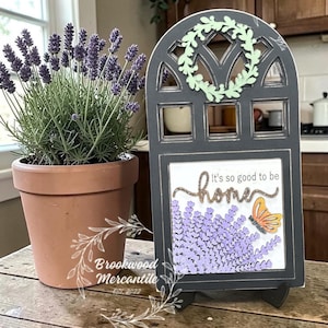 May include: A decorative wooden sign with a black frame and a light green wreath at the top. The sign features the text "It's so good to be home" above a lavender and butterfly design. A potted lavender plant sits to the left.