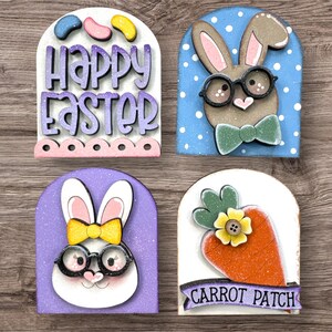 Pode incluir: Quatro decora&ccedil;&otilde;es de P&aacute;scoa em madeira. Uma diz "Happy Easter" com gomas. Outra apresenta um coelho com &oacute;culos e gravata borboleta sobre um fundo azul. Uma terceira mostra um coelho com &oacute;culos e la&ccedil;o amarelo. A &uacute;ltima diz "Carrot Patch".