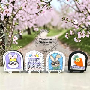 May include: Four Easter-themed wooden decorations. Each features a different design: a bunny with glasses, the text "Happy Easter", a bunny with glasses and a bow tie, and a carrot patch. The decorations are in front of a blurred floral background.