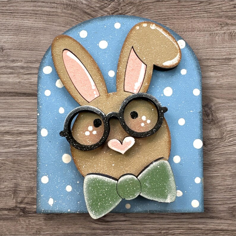 Pode incluir: Decora&ccedil;&atilde;o de coelho da P&aacute;scoa em madeira com rosto e orelhas castanhas claras e gravata borboleta verde. O coelho usa &oacute;culos pretos e tem um nariz em forma de cora&ccedil;&atilde;o rosa. O fundo &eacute; azul claro com bolinhas brancas.