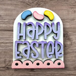 Pode incluir: Decora&ccedil;&atilde;o de P&aacute;scoa em madeira com as palavras "Happy Easter" em letras roxas e brancas com glitter. A decora&ccedil;&atilde;o tem tr&ecirc;s formas de gomas coloridas no topo e uma borda rosa com vieiras na parte inferior. O fundo &eacute; verde claro.