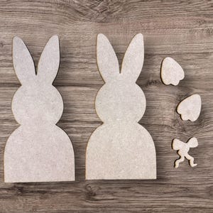 May include: Two wooden bunny cutouts and three smaller wooden shapes, including two ear pieces and a bow, are arranged on a wood-grain surface. The bunny cutouts are light gray and have a simple, minimalist design.