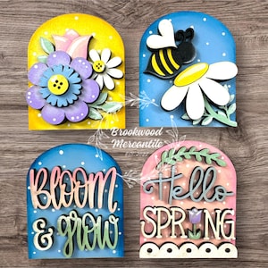 May include: Four wooden spring-themed decorations. One features a purple flower, another a bee and daisy, a third says "Bloom & Grow", and the last says "Hello Spring". Each has a colorful arched background and white polka dots.