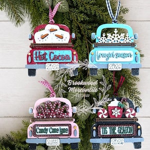 May include: Four wooden truck ornaments with hanging ribbons. The red truck features snowmen and says Hot Cocoa 2026. The blue truck has snowflakes and says freezin season 2026. The pink truck has candy canes and says Candy Cane lane SWEET. The black truck has cocoa and says TIS THE SEASON 2026.