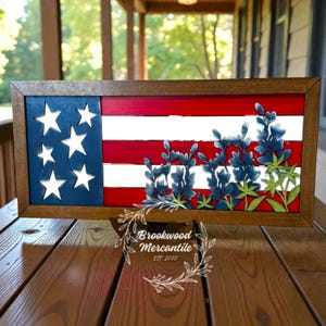 May include: Wooden framed artwork depicting the American flag with bluebonnet flowers. The flag features a blue field with white stars and red and white stripes. Bluebonnet flowers are layered over the stripes. The frame is brown, and the artwork is displayed on a wooden surface.