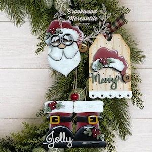 May include: Three wooden Christmas ornaments featuring a Santa face with glasses, a tag with a red hat and the word Merry, and a pair of boots with the word Jolly. Each piece includes painted details like holly berries, buttons, and plaid ribbon.