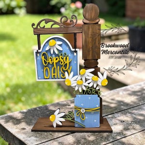 May include: Wooden sign with a blue background and the words "Dopsy Daisy" in yellow script. The sign features a decorative wooden post with a jar of white daisies with yellow centers. The sign is on a wooden base.