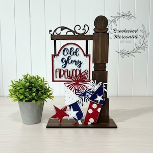 May include: A wooden patriotic sign with the words "Old Glory Fireworks" in blue and red. The sign features a dark brown post with decorative fireworks and a star. A small potted plant is on the left.