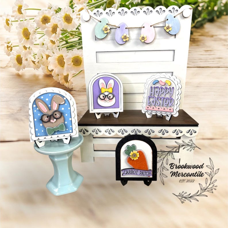 Pode incluir: Uma exibi&ccedil;&atilde;o de P&aacute;scoa em madeira branca com sinais de coelhinho e cenoura em miniatura. Os sinais apresentam v&aacute;rios desenhos, incluindo um coelhinho com &oacute;culos e as palavras "Happy Easter". Uma grinalda de ovos de P&aacute;scoa est&aacute; pendurada acima.