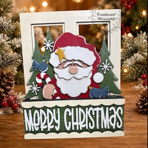 May include: A wooden window-shaped Christmas decoration featuring a layered Santa Claus with a candy cane, green pine trees, blue birds, and white snowflakes. The base is a green planter box with the white text Merry Christmas printed on it.
