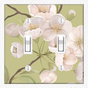 May include: A double light switch cover featuring a floral design. The cover has a light green background with a pattern of white and pink blossoms and buds. The switches are white.