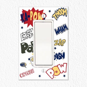 May include: White light switch cover with comic book style sound effects in various colors. The cover features words like "KA-POW," "CRASH," and "ZAP" in bold, stylized fonts. The switch opening is rectangular.