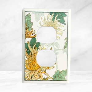May include: A white electrical outlet cover featuring a floral design with yellow and green chrysanthemums. The flowers are set against a cream background with a green border. The cover has two circular openings for the outlets.