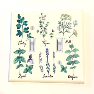 May include: A decorative light switch cover featuring watercolor illustrations of herbs including parsley, thyme, dill, basil, lavender, and oregano. The names of the herbs are written below each illustration. The cover has two light switches.