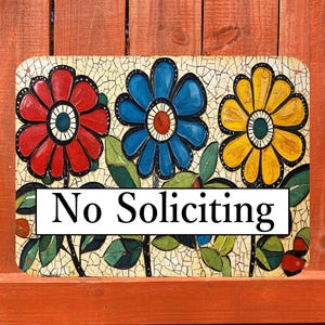 May include: Rectangular ceramic sign with three colorful flowers: red, blue, and yellow. The flowers have black outlines and are set against a mosaic background. The sign reads "No Soliciting" in a white rectangle.