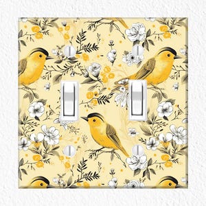 May include: A decorative light switch cover featuring a yellow background with a floral and bird design. The design includes yellow birds, white flowers, and black leaves. The cover has two white light switches.