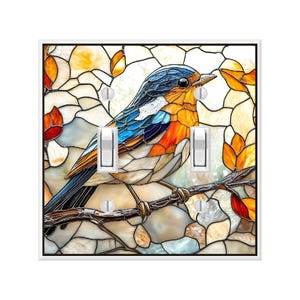 May include: A decorative light switch cover featuring a stained-glass style bird design. The bird has blue, orange, and white feathers, perched on a brown branch. The background has a mosaic of beige, orange, and white shapes.