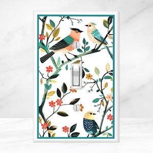 May include: A decorative light switch cover featuring a colorful illustration of three birds perched on branches with leaves and flowers. The birds have orange, blue, and yellow plumage. The cover has a white background and a teal border.