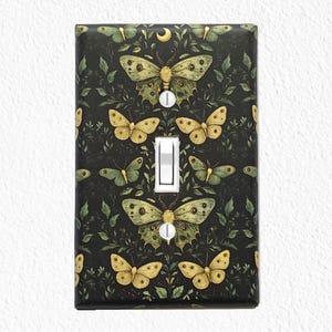 May include: A decorative light switch cover featuring a dark background with a pattern of butterflies and foliage in shades of green and yellow. The switch itself is white. The design includes a crescent moon.