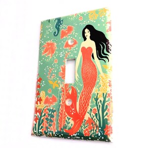 May include: A light switch cover with a mermaid illustration. The mermaid has long black hair, a red top, and a coral-colored tail. The background is a green and blue ocean scene with coral, seahorses, and fish.