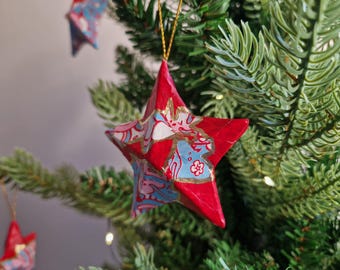 May include: A five-pointed star ornament featuring a vibrant red base with light blue and white floral patterns. The star has gold-colored painted accents along the edges and is suspended by a thin gold string for hanging on a tree branch.