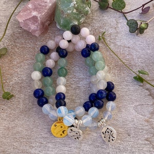 May include: Three layered bracelets with blue, green, and white beads. The bracelets have gold and silver charms with celestial symbols.