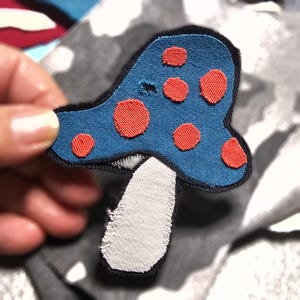 Può includere: Una toppa a forma di fungo blu con pois rossi e gambo bianco. La toppa ha un bordo nero ed è realizzata in tessuto. Il fungo è un design stravagante, adatto per abbigliamento o accessori.
