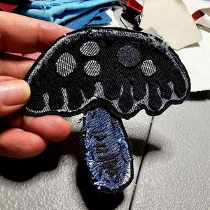 May include: A black mushroom-shaped patch with white polka dots and a blue denim stem. The patch is made of fabric and has a stitched border. The mushroom cap has a textured appearance.