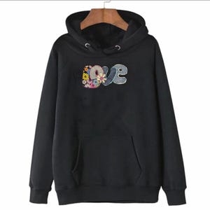 May include: Black hooded sweatshirt with a front pocket and drawstring hood. The word "love" is embroidered on the front in a floral and gray design. The sweatshirt is hanging on a wooden hanger.
