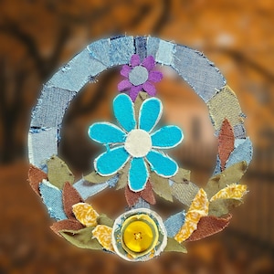Peut inclure: Une couronne de signe de paix faite à la main, avec une base en denim, ornée d'une grande fleur turquoise, d'une petite fleur violette et de feuilles en tissu. Un bouton jaune est en bas. Un article décoratif coloré.